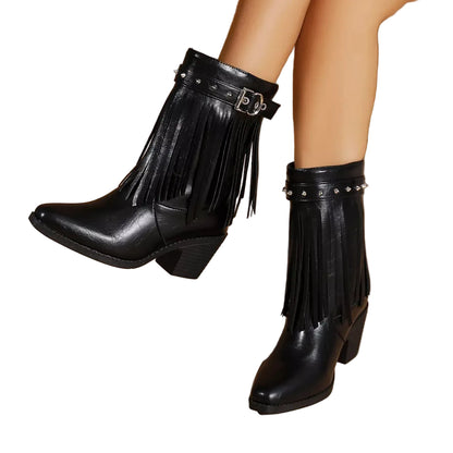 A pair of black faux-leather ankle boots with fringe detailing, a studded strap, and a low block heel, shown on a model's feet against a beige background.