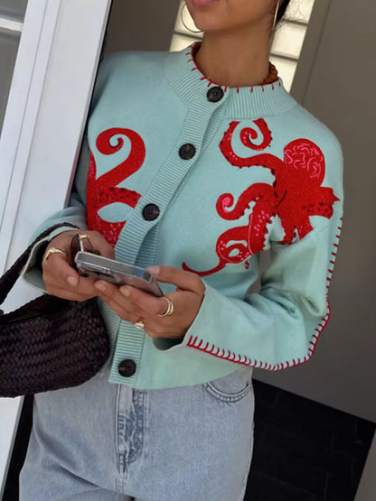 A woman wearing a mint green knit cardigan with large red octopus motifs and red contrast stitching, paired with light blue jeans and holding a phone.