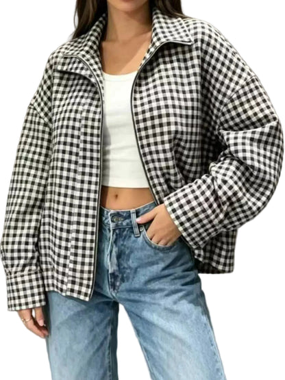 A woman wearing a black and white gingham zip-up jacket over a white crop top and light blue jeans, standing against a neutral gray wall.
