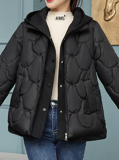 A woman wearing a black quilted puffer jacket with a hood, layered over a cream turtleneck and blue jeans, shown from the neck down against a blue paneled wall.