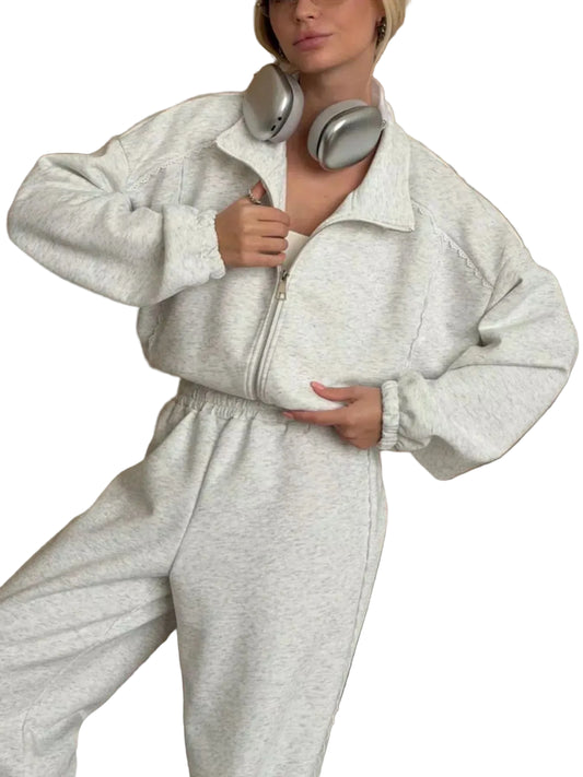 A woman wearing a light heather gray zip-up sweatshirt and matching sweatpants, with silver headphones around her neck, posed in front of a wood-paneled wall.