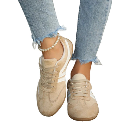 A person wearing beige suede sneakers with white side stripes, paired with frayed light blue jeans and a pearl ankle bracelet, standing on a wooden deck.