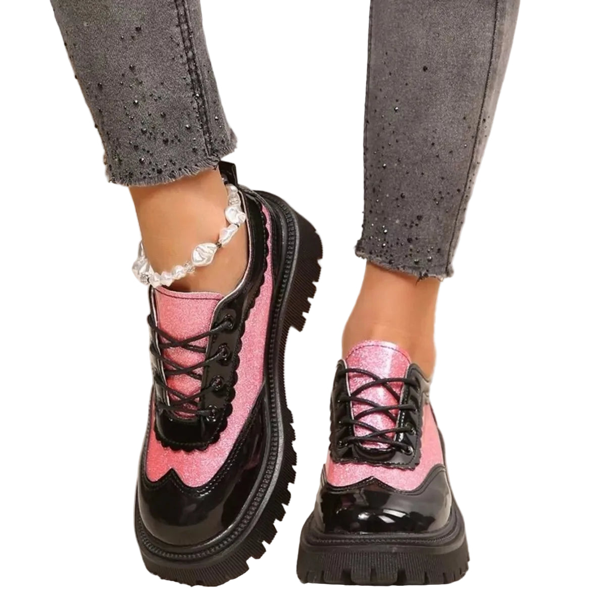 A pair of chunky lace-up platform sneakers in glossy black and pink glitter, shown on a model's feet wearing gray jeans and a pearl anklet.