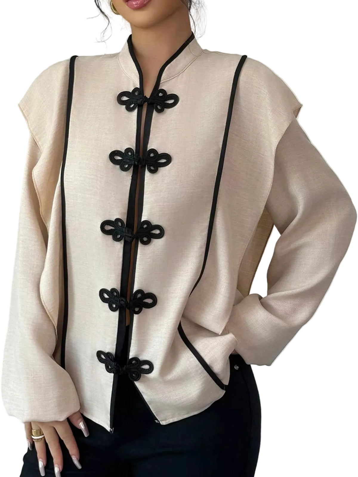 A woman wearing a beige long-sleeve blouse with black trim and decorative black frog closures down the front, paired with black pants, against a plain light gray background.