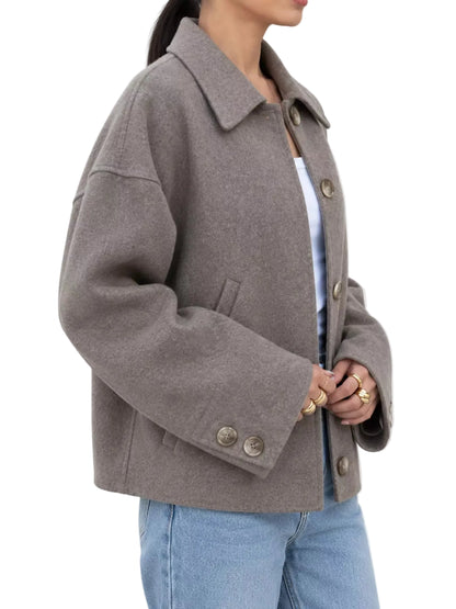 A woman wearing a taupe-brown oversized button-front coat with a collar, paired with a white top and light blue jeans, shown from the side against a plain white background.