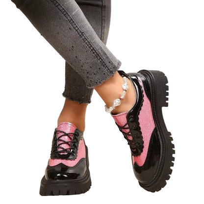 A person wearing chunky platform lace-up shoes with glossy black and pink panels, paired with gray jeans and a silver anklet, in a cozy indoor setting.