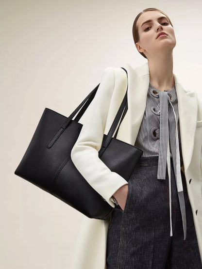 A woman wearing a white blazer, striped blouse, and dark denim skirt carries a large black leather tote bag over her shoulder against a plain light beige studio background.