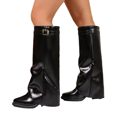 A pair of black faux-leather knee-high boots with a buckle detail on the side, shown on a model's feet in an indoor setting.