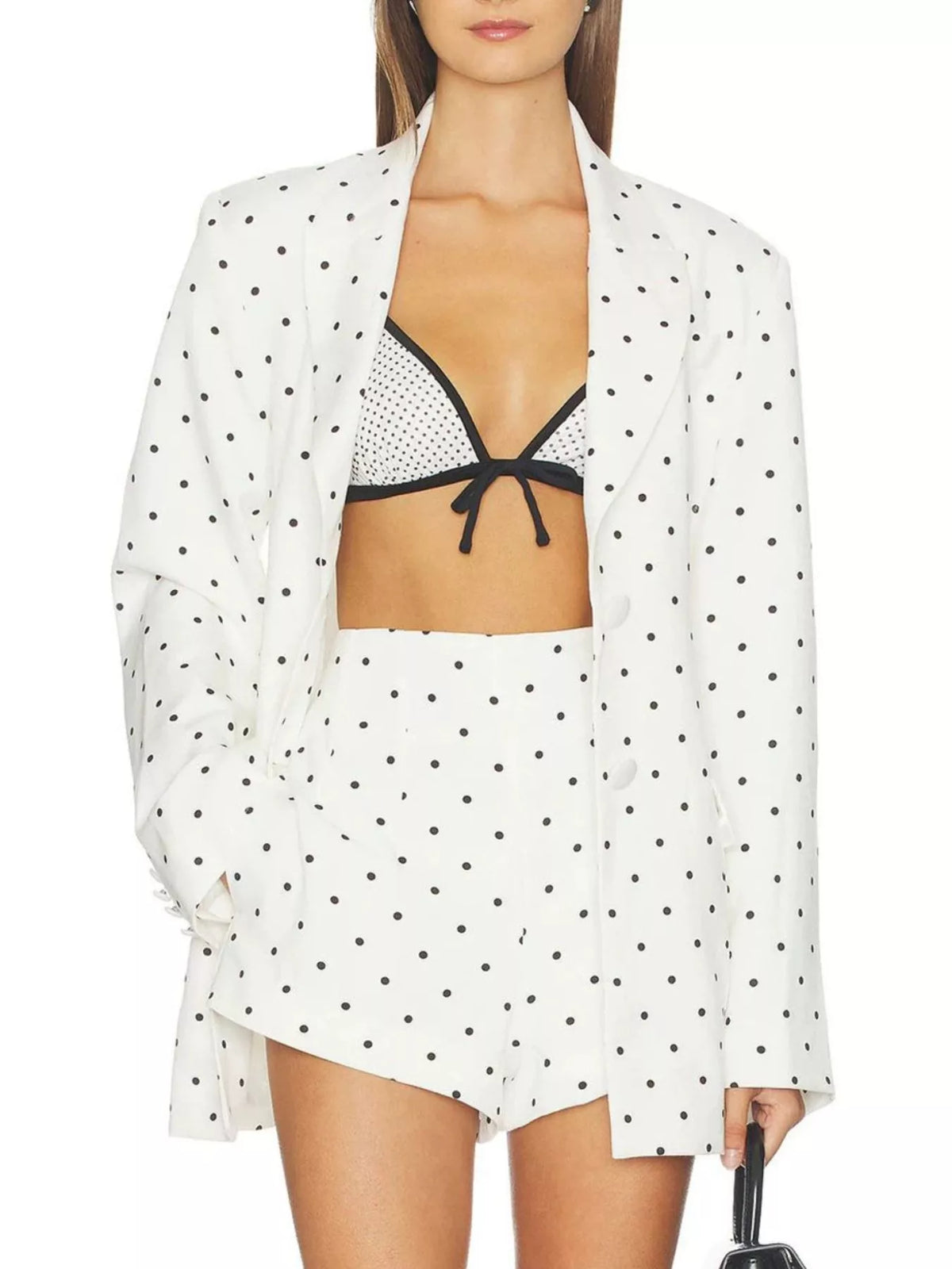 A model wearing a white two-piece set with black polka dots, consisting of a long-sleeve open-front jacket and matching shorts, styled over a black and white polka-dot bikini top.