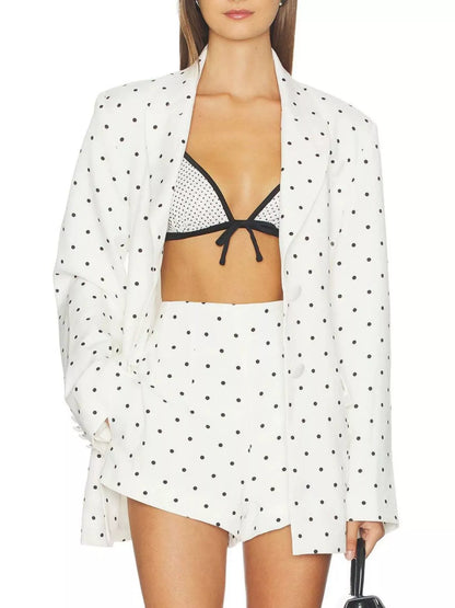 A model wearing a white two-piece set with black polka dots, consisting of a long-sleeve open-front jacket and matching shorts, styled over a black and white polka-dot bikini top.