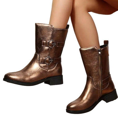 Metallic bronze ankle boots with buckle straps and a side zipper, shown on a model's feet resting on a wooden stool.