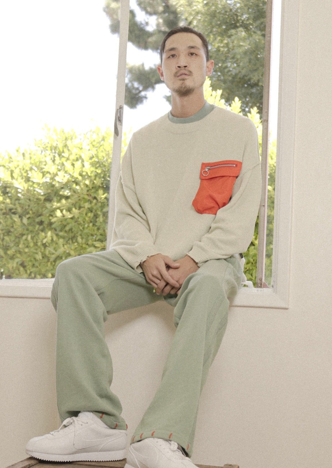 Man sitting on a windowsill wearing a light gray sweater with a red pocket and green pants.