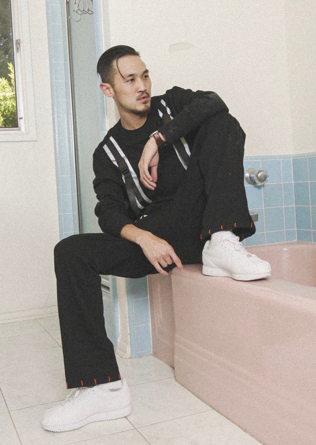Person wearing a black outfit with white accents sitting on a pink bathtub in a bathroom.