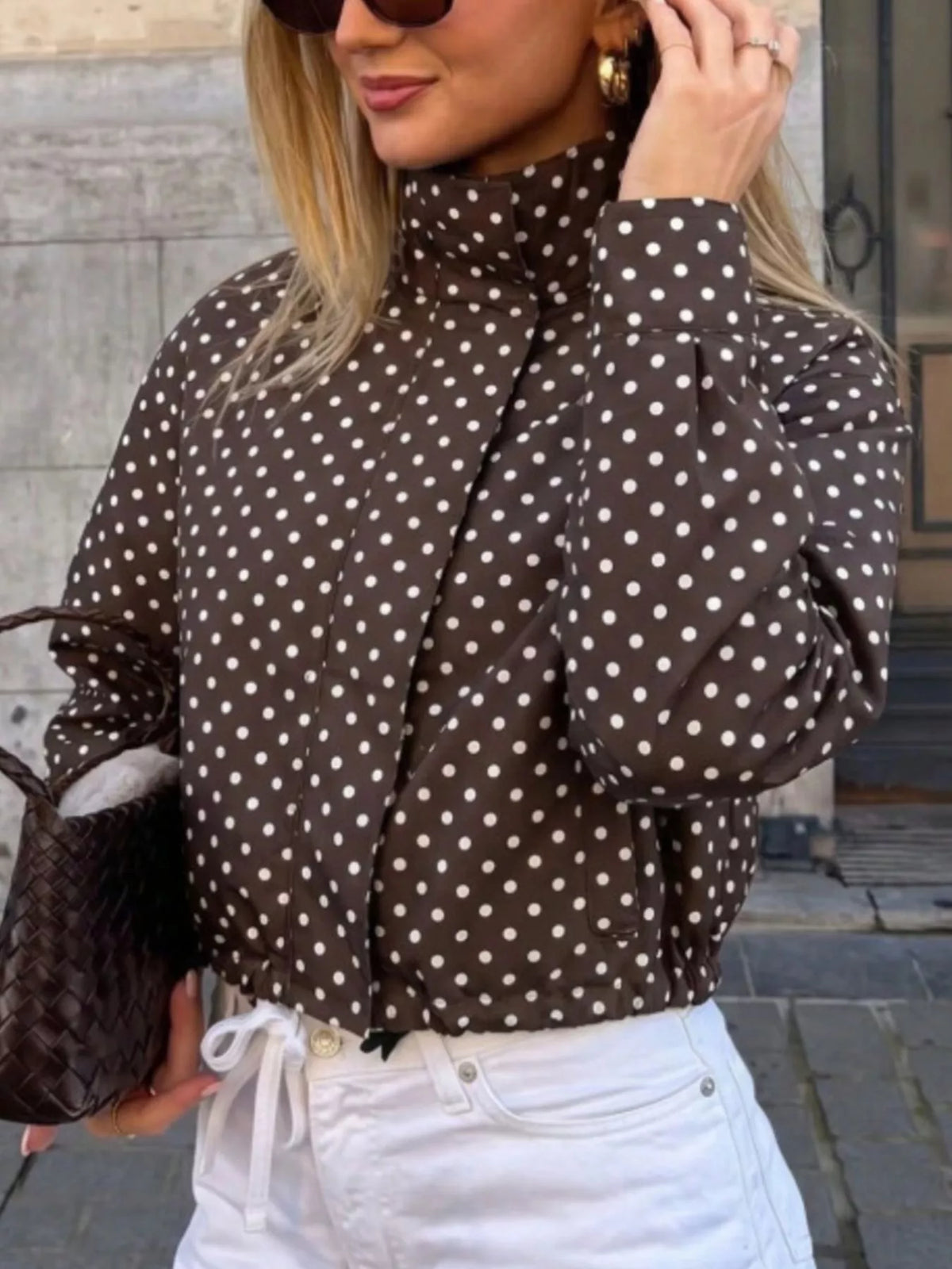 A woman wearing a brown long-sleeve mock-neck blouse with white polka dots, paired with white drawstring jeans and holding a woven brown handbag.