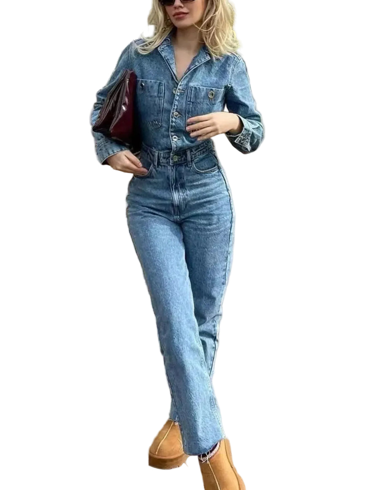 A woman wearing a light-wash blue denim jumpsuit with a button-up shirt collar and long sleeves, paired with tan boots and carrying a dark red bag, standing on a wooden deck.