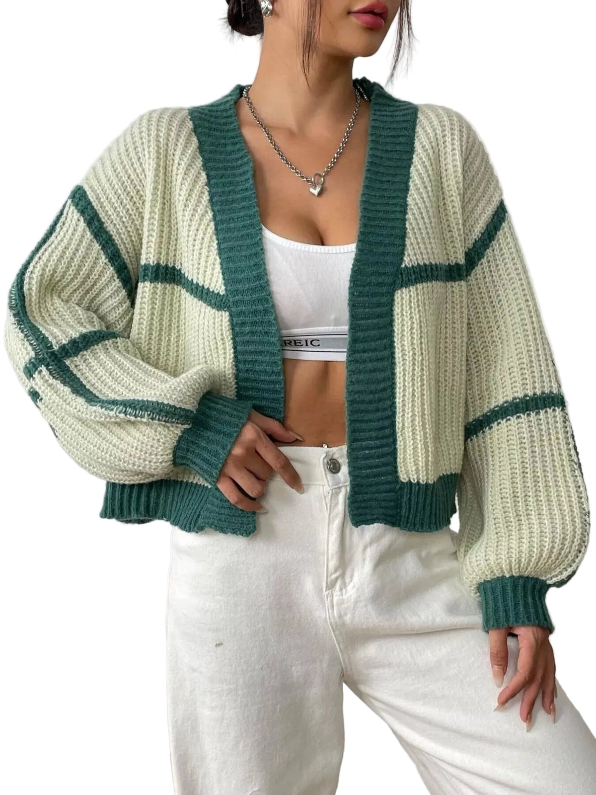 A woman wearing a chunky knit cardigan with a cream and dark green color-blocked pattern, styled over a white crop top and white jeans.