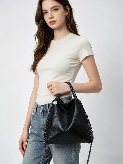 A woman wearing a cream short-sleeve t-shirt and light blue jeans carries a black leather bucket bag with a silver-tone chain strap against a light gray studio background.