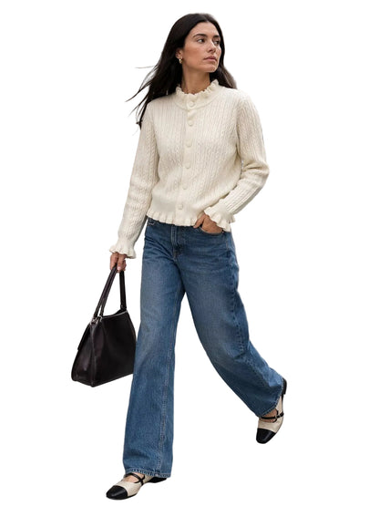 A woman walking on a city street crosswalk wearing a cream cable-knit mock-neck cardigan, wide-leg blue jeans, and black-and-white shoes, holding a dark brown leather handbag.