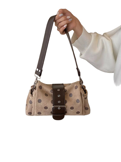 A person holds up a beige shoulder bag with dark brown polka dots and a dark brown leather strap and front buckle detail, set against an indoor background.