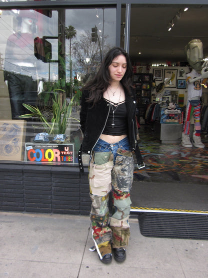 A woman stands on a sidewalk in front of a clothing store window, wearing a black zip-up jacket, a black crop top, blue jeans, and highly detailed, multi-colored patchwork cargo pants with camouflage and floral patterns.
