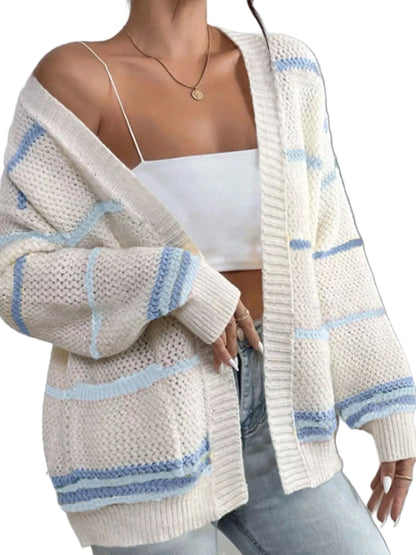 A woman wearing a chunky knit open-front cardigan in off-white with light blue stripes, layered over a white camisole and light-wash jeans.