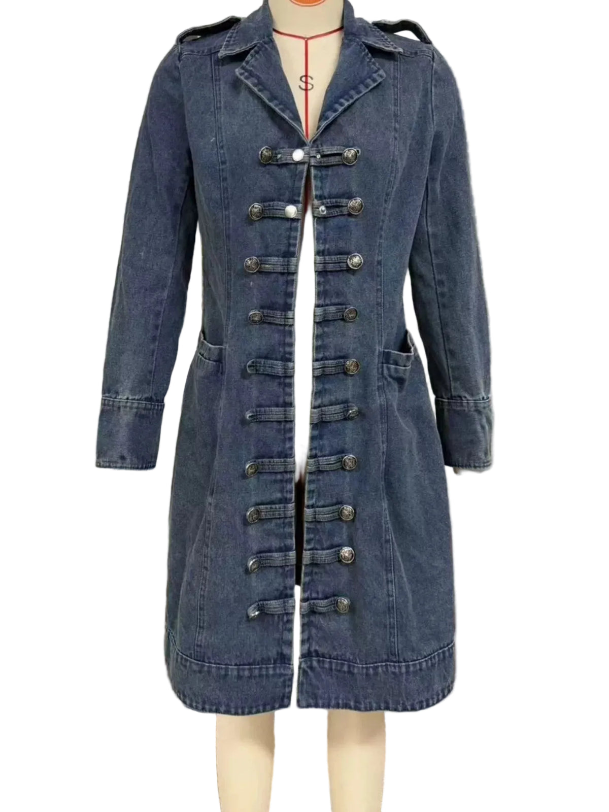 A blue denim military-style coat displayed on a mannequin, featuring a front zipper, decorative metal buttons, and shoulder epaulets, set against a window with horizontal blinds.