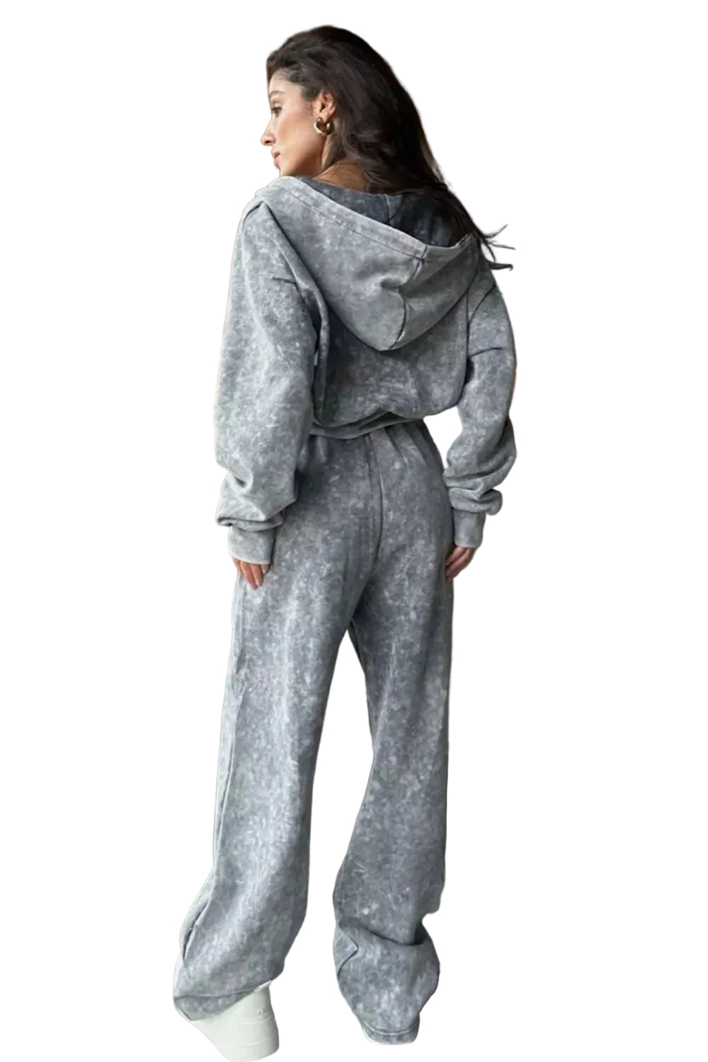 A woman wearing a matching gray hooded sweatshirt and wide-leg sweatpants set, standing in a modern living room with a fireplace and sofa in the background.
