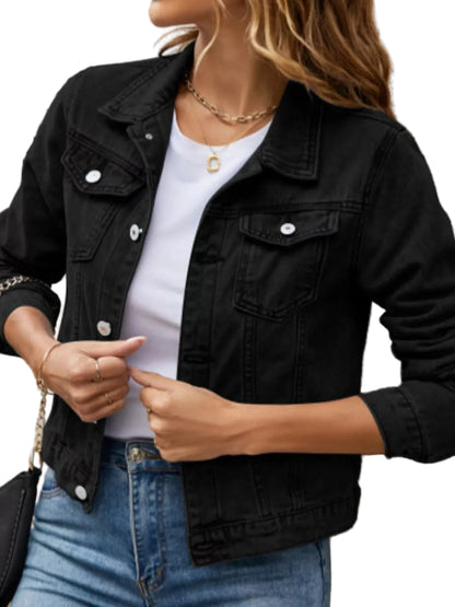 A woman wearing a black denim jacket over a white top and blue jeans, standing against a stone wall.