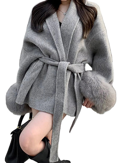 A woman wearing a light gray belted coat with fluffy faux-fur trim at the cuffs and hem, standing outdoors next to a gray pole on a street.