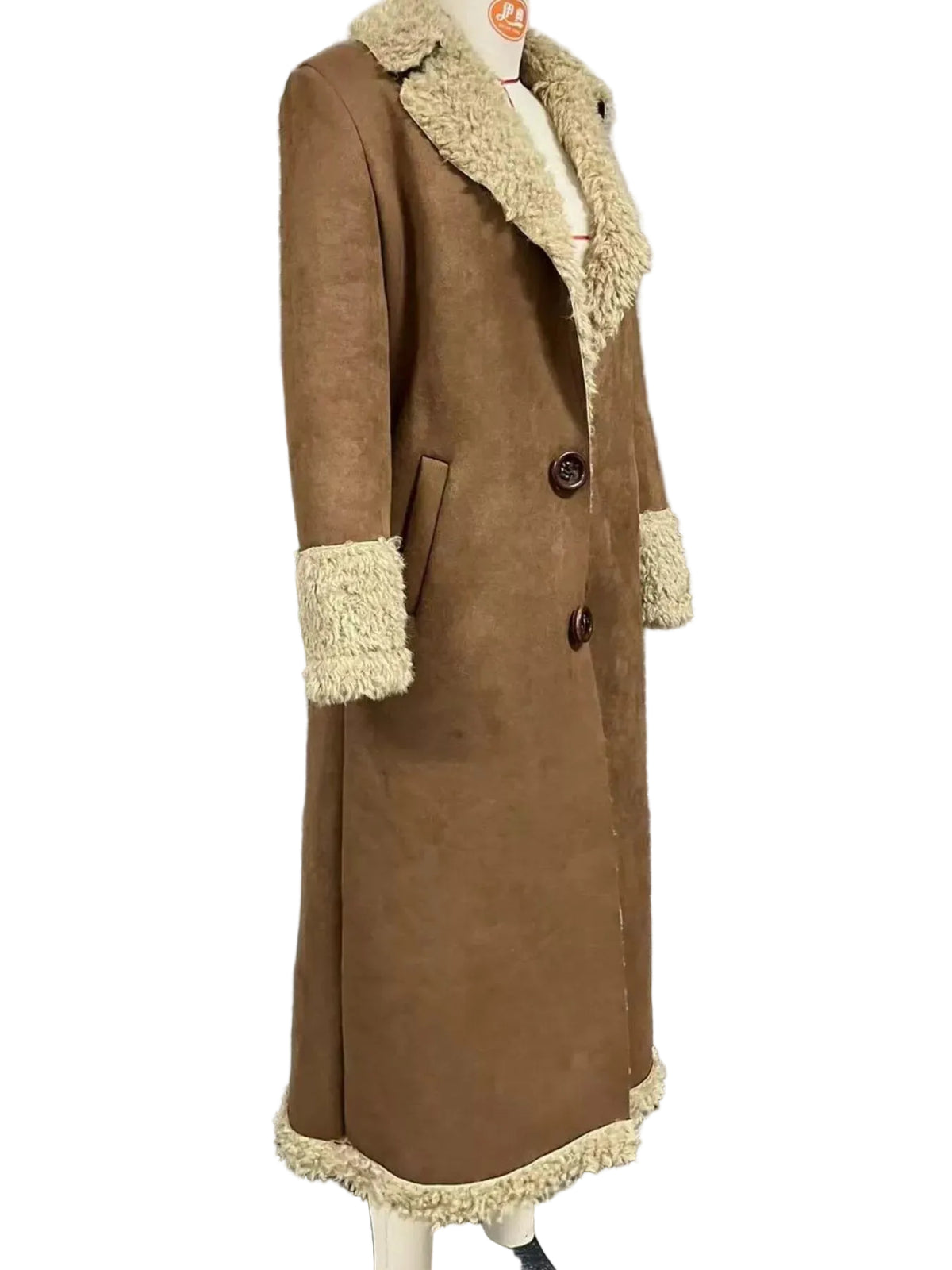 A long brown suede-like coat with cream shearling-style collar, cuffs, and hem, displayed on a mannequin in front of a window with horizontal blinds.