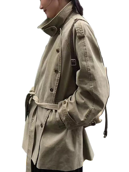 A person wearing a light khaki trench coat with a belted waist, shown in profile outdoors next to a black car.