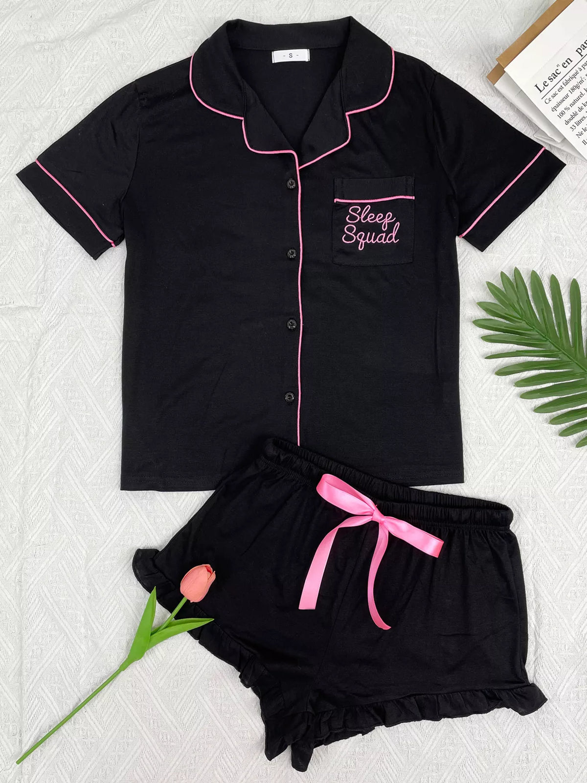 A two-piece women's pajama set laid flat on a light textured background, featuring a short-sleeve button-front top and matching shorts. The pajamas are black with pink piping and a pink drawstring, with 'Sleep Squad' embroidered on the chest pocket.