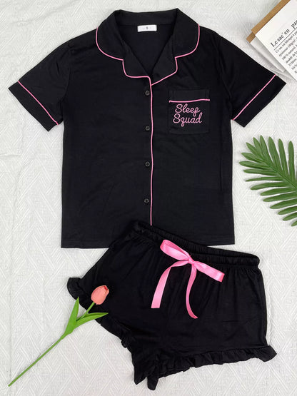 A two-piece women's pajama set laid flat on a light textured background, featuring a short-sleeve button-front top and matching shorts. The pajamas are black with pink piping and a pink drawstring, with 'Sleep Squad' embroidered on the chest pocket.