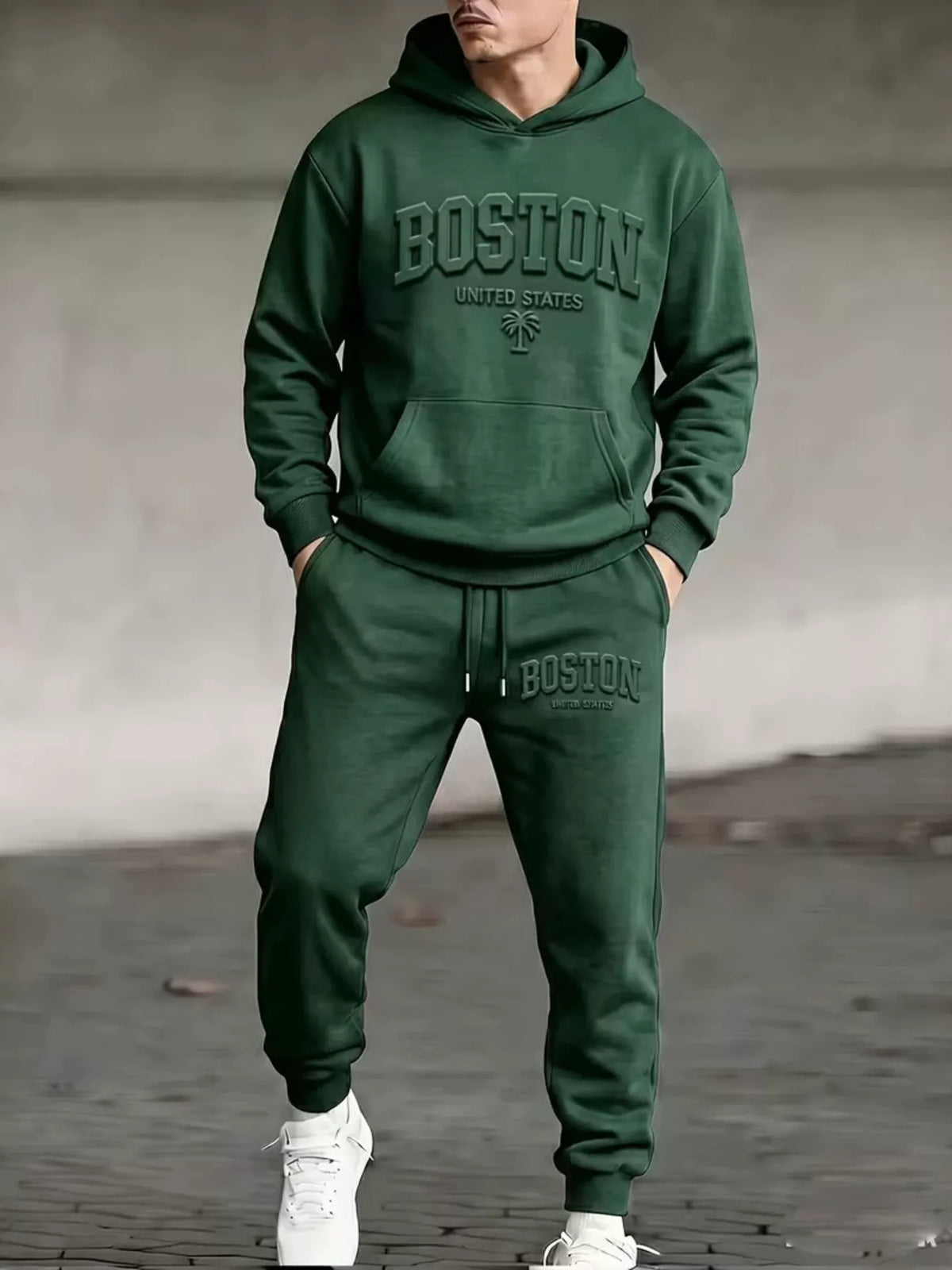 A man wearing a dark green matching hoodie and sweatpants set with 'BOSTON UNITED STATES' printed on the front and left thigh, paired with white sneakers, standing against a concrete wall.