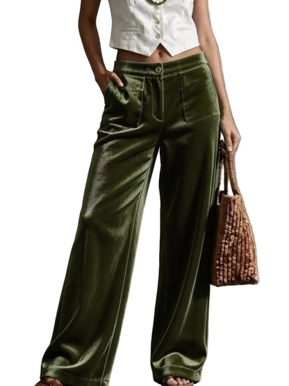 A model wearing high-waisted, wide-leg olive green velvet pants, styled with a white cropped vest, holding a brown woven handbag against a textured gray concrete wall.