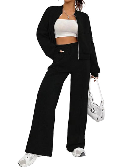 A model wearing a black two-piece lounge set consisting of a cropped zip-up jacket and matching wide-leg pants, styled with a white crop top, white sneakers, and a white handbag against a textured gray wall.