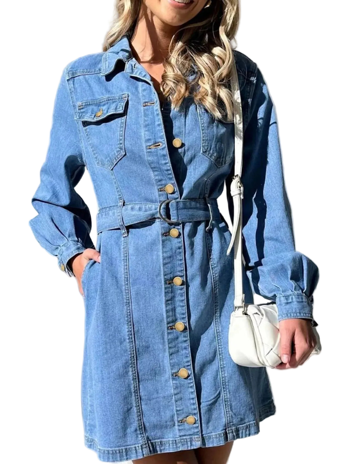 A woman wearing a light blue denim shirt dress with a button-down front, chest pockets, and a matching belt. She is holding a small white shoulder bag against a background of white curtains.
