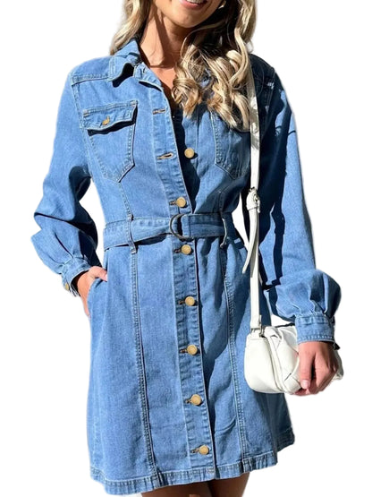 A woman wearing a light blue denim shirt dress with a button-down front, chest pockets, and a matching belt. She is holding a small white shoulder bag against a background of white curtains.