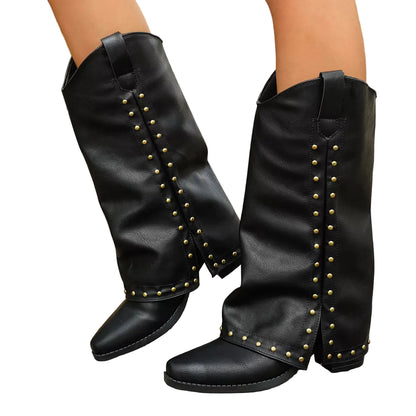 A pair of black mid-calf boots with gold-tone metal studs, shown on a person's feet standing on a wooden boardwalk outdoors.
