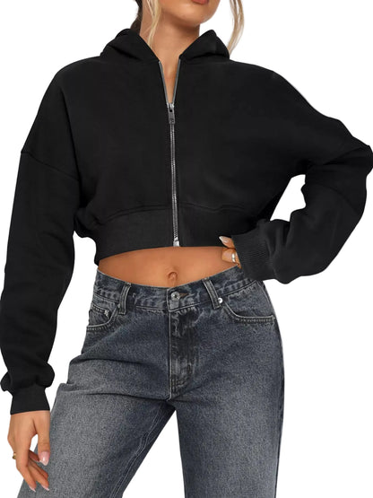 A woman wearing a black cropped zip-up hoodie with a silver zipper and dark gray denim jeans, shown from the neck down against a light gray studio background.