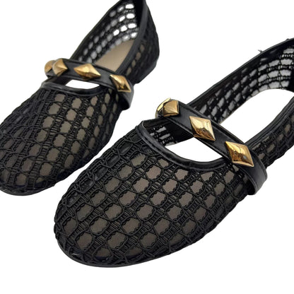 A pair of black crochet mesh Mary Jane flats with glossy black straps and gold pyramid stud embellishments, shown on a light gray background.