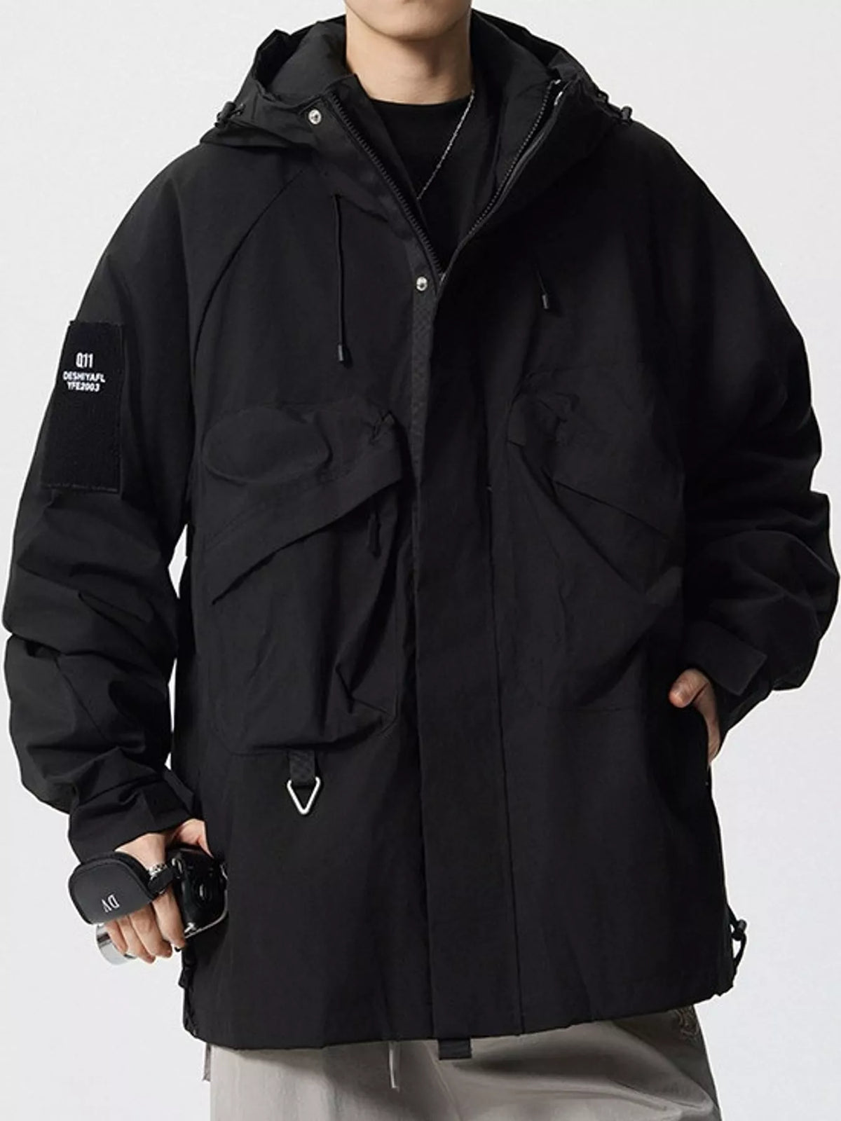 A person wearing an oversized black hooded jacket with multiple front pockets, a black shirt underneath, and light gray pants, photographed against a plain light background.
