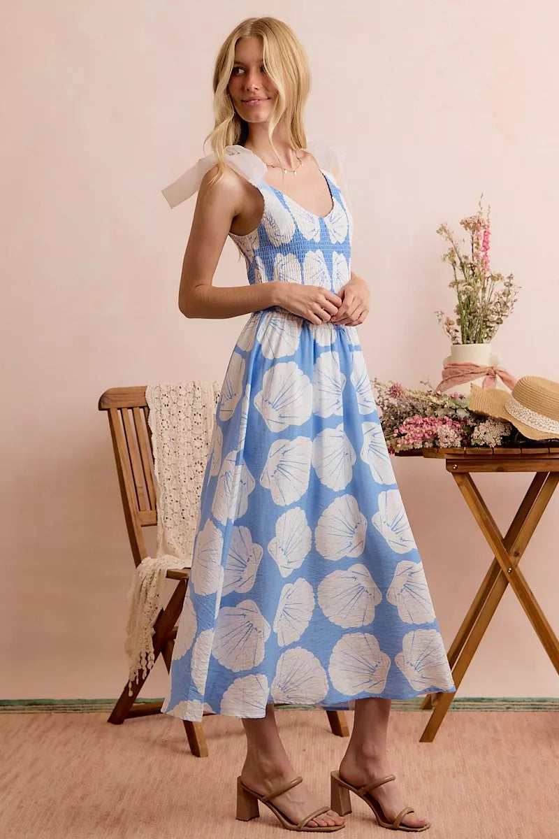 Woman wearing a blue floral dress standing in a room with a wooden table and chair.