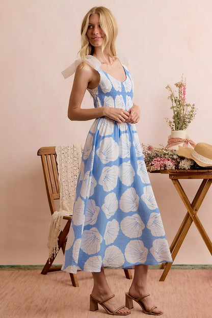 Woman wearing a blue floral dress standing in a room with a wooden table and chair.
