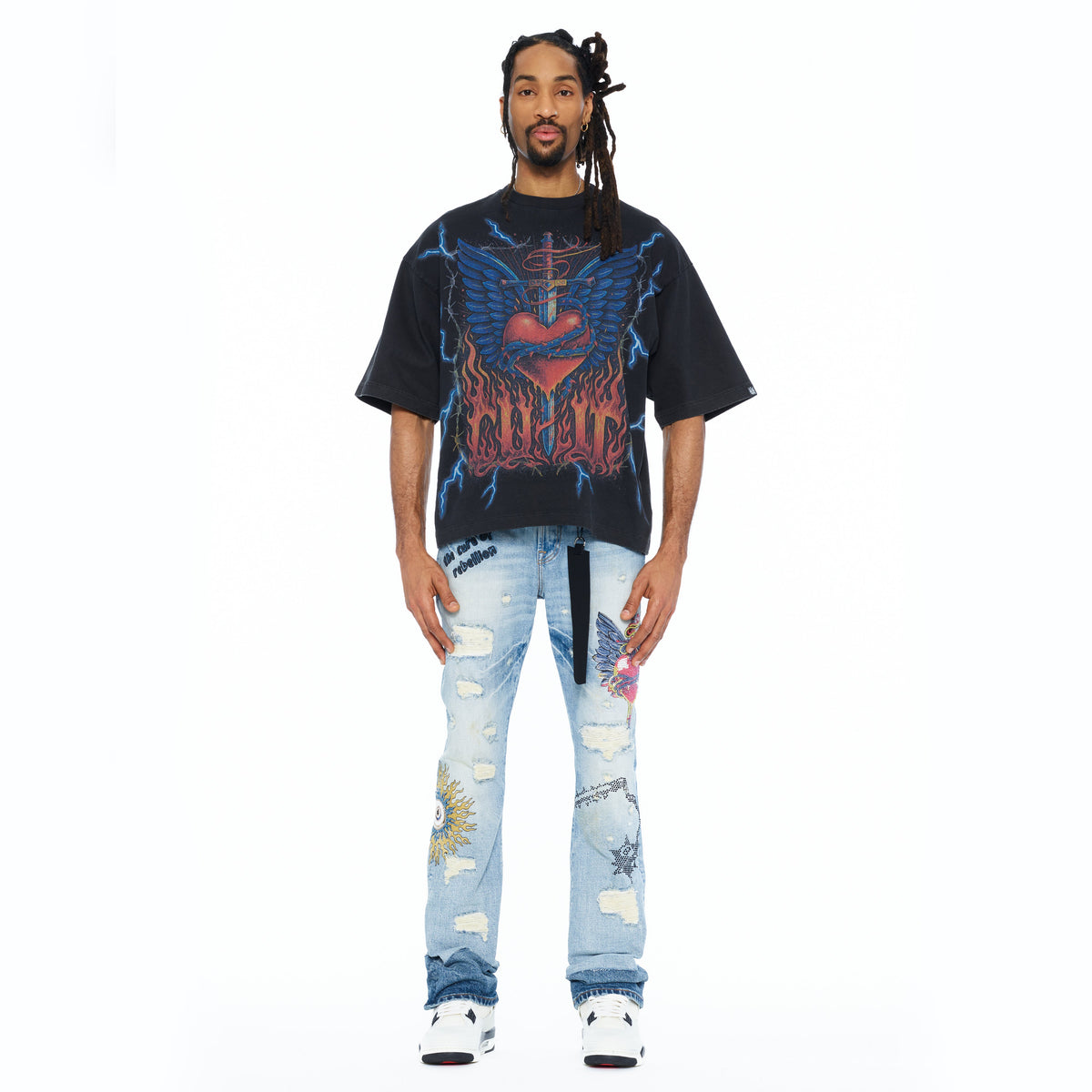 Man wearing a black graphic t-shirt and light blue jeans with patches on a white background