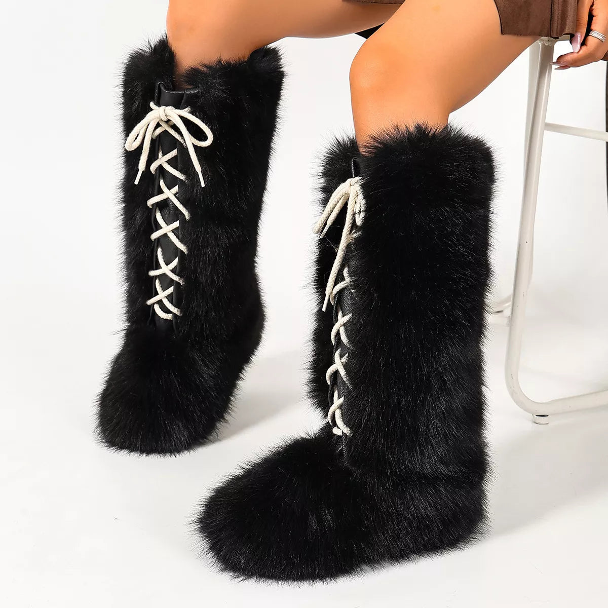 A pair of black faux-fur knee-high boots with white lace-up front details, shown on a model's legs against a white studio background.