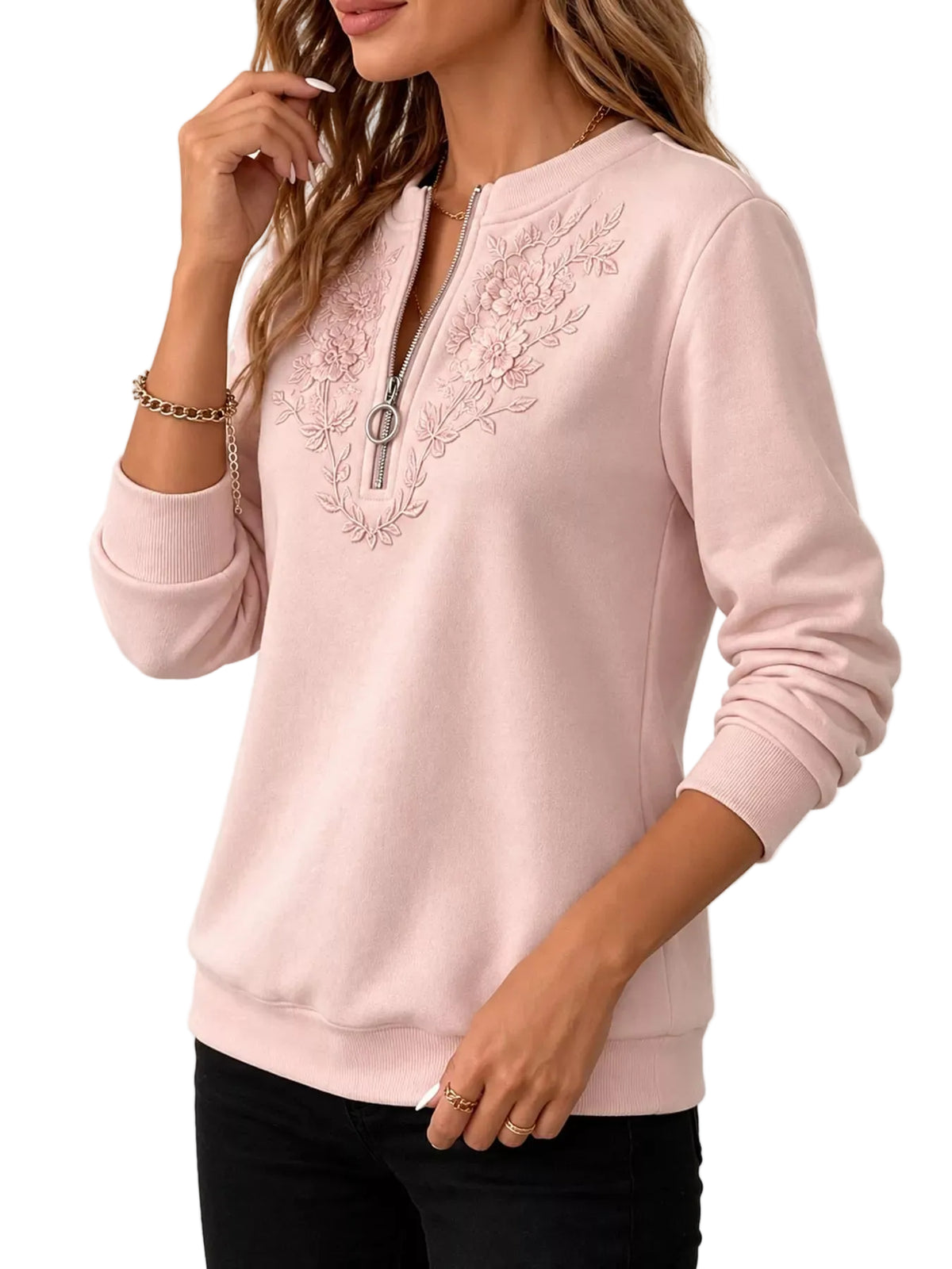 A woman wearing a light pink long-sleeve sweatshirt with a half-zip neckline and floral embroidery on the chest, paired with black pants against a white background.