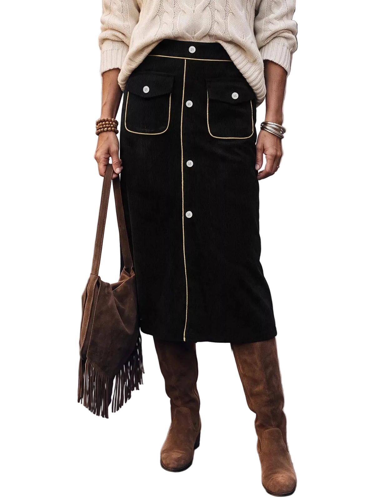 A person standing on a city street wearing a black midi skirt with gold trim and white buttons, a cream cable-knit sweater, brown suede knee-high boots, and carrying a brown fringe shoulder bag.