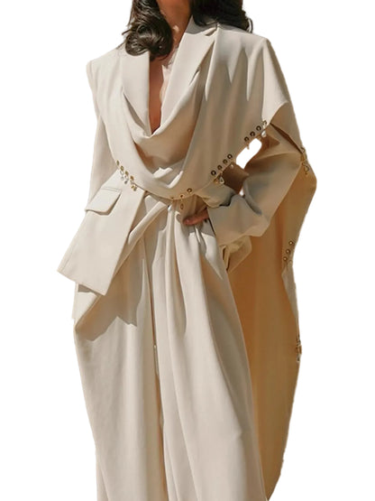 A model wearing a cream-colored, oversized draped coat with structured shoulders, a matching tie belt, and decorative gold-tone studs along the sleeve edges, set against a beige stone wall background.