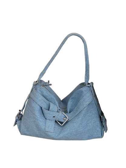 A light blue denim shoulder bag with a front flap and buckle detail, resting on a wooden floor next to a bed with white bedding.